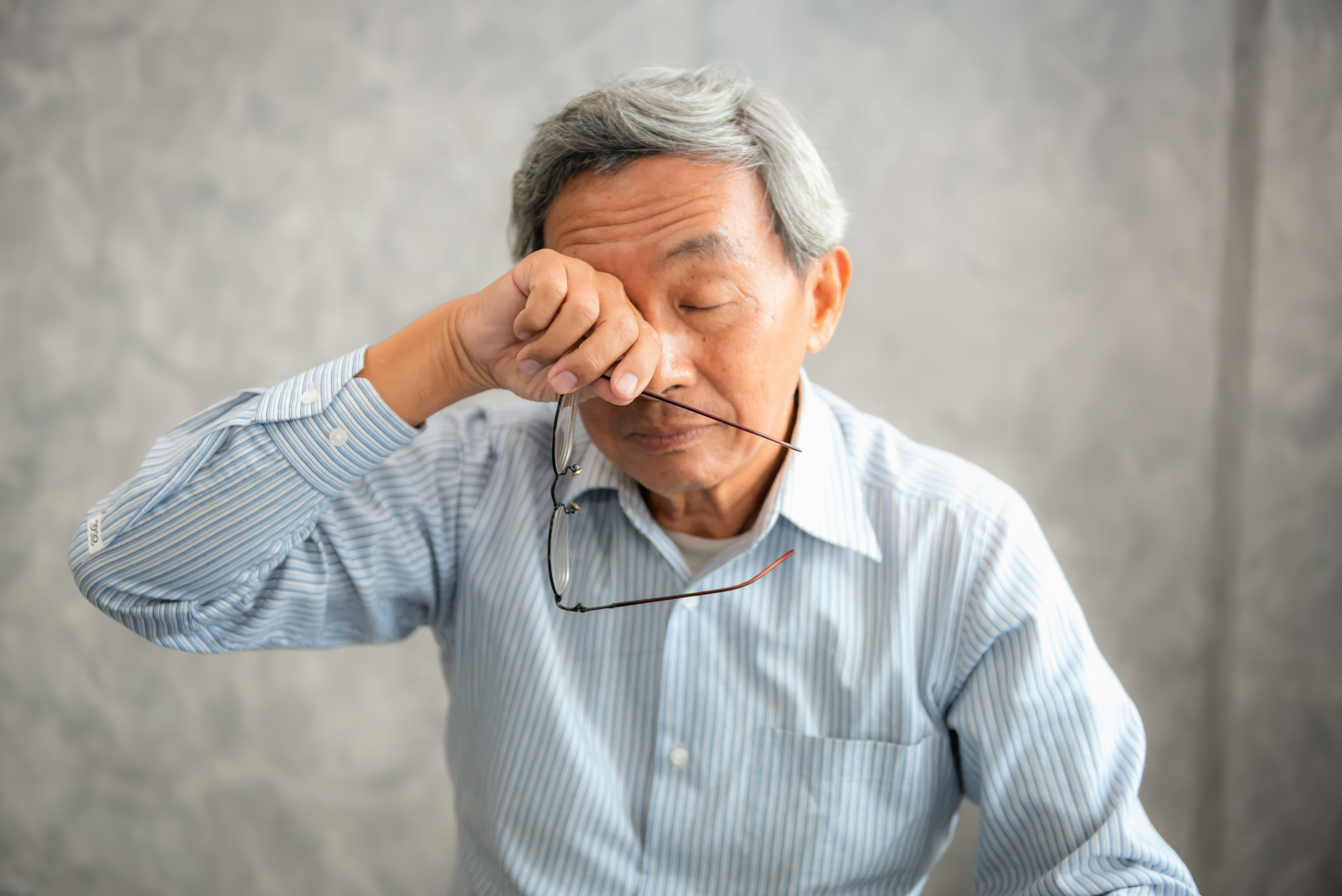 An older man holds his glasses in his hand and rubs his eyes. He appears to be fatigued. 